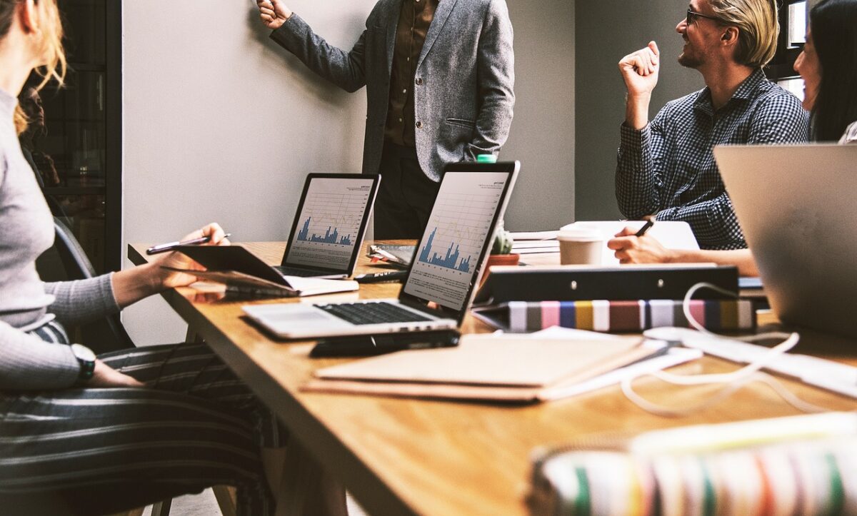 A professional office with three people talking. Two are at the conference table with laptops and the third is at the whiteboard.